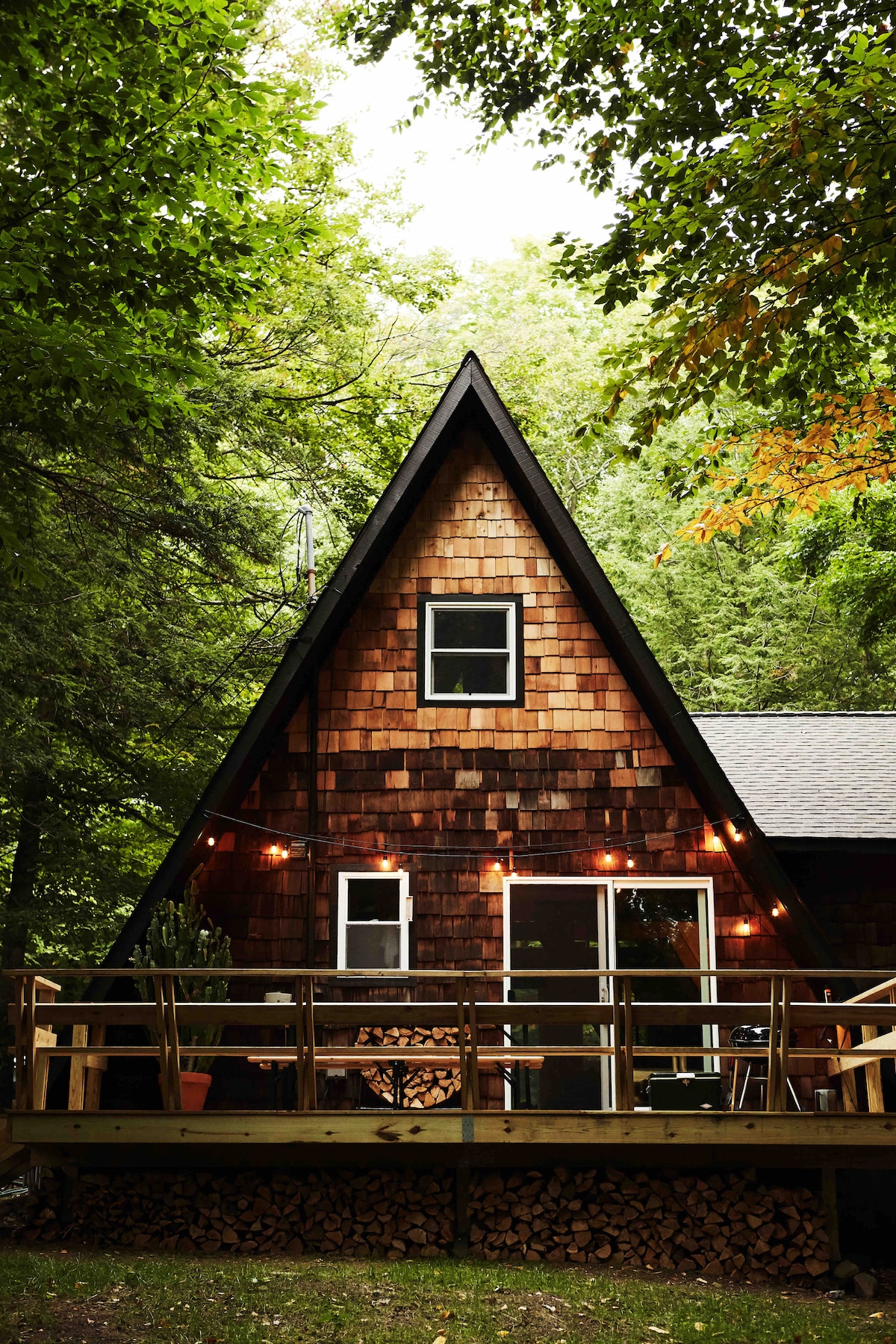 A charming cabin with an A-frame design is surrounded by lush green trees. The exterior features wooden shingles and large windows. A spacious porch displays stacked firewood and is adorned with warm string lights, providing a cozy touch to the inviting outdoor space.