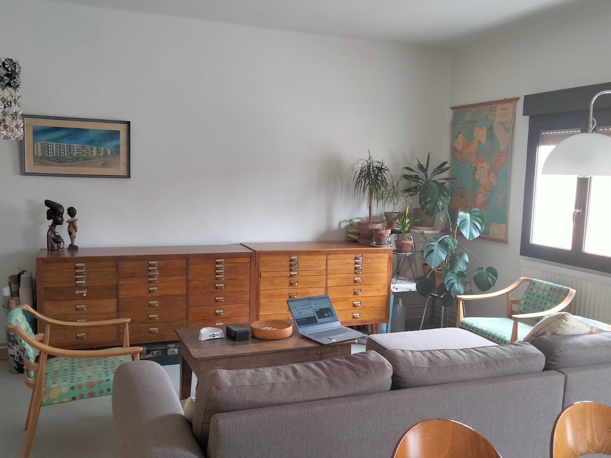 A spacious living area is visible, featuring a comfortable sofa and two accent chairs. Natural light enters through a window, illuminating a wooden cabinet and a wall map. Decorative plants add a touch of greenery to the inviting ambiance.