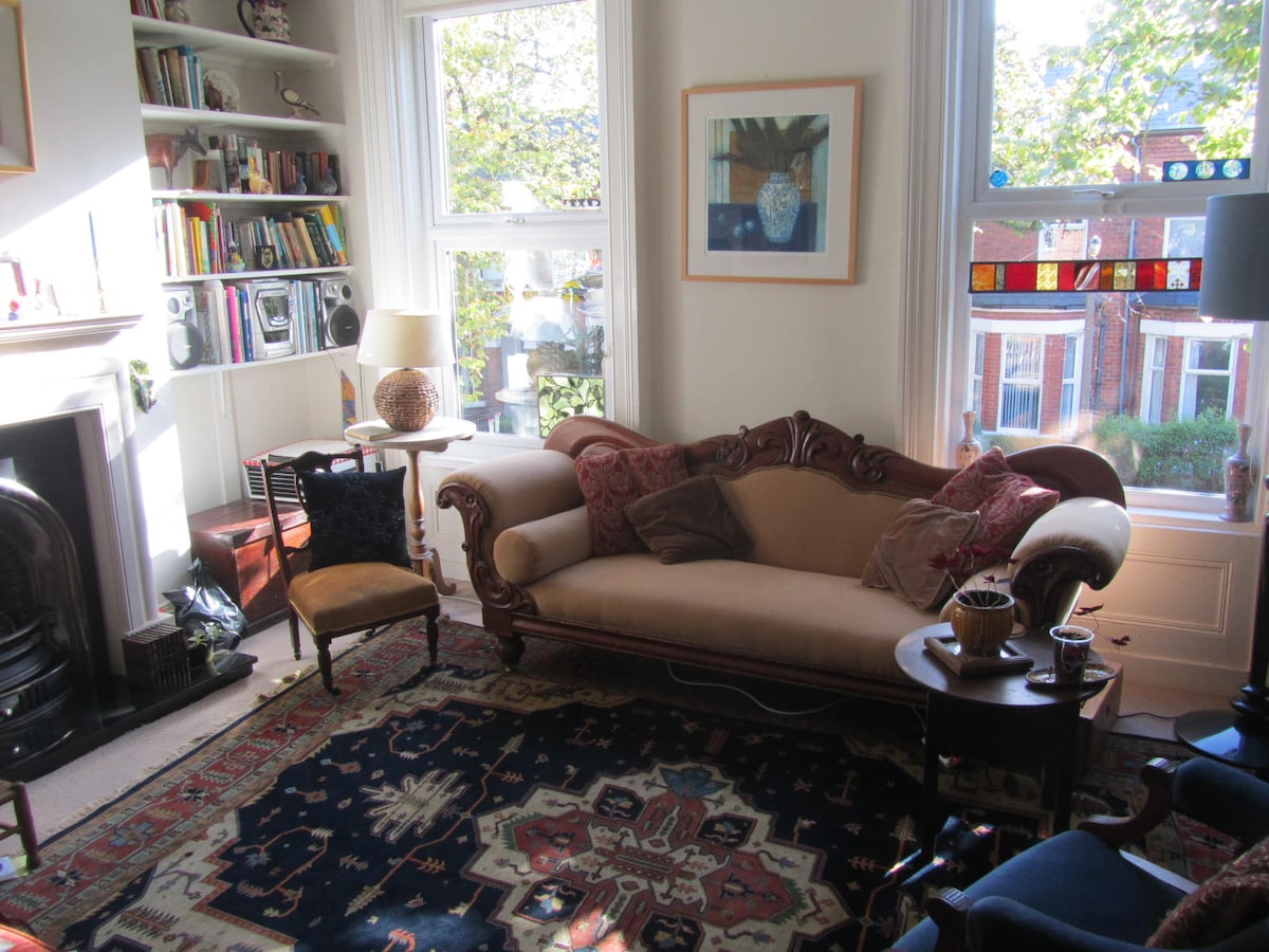 A bright sitting room is filled with natural light from large windows, showcasing a mix of books and art on shelves. A plush, upholstered couch and a dark wooden table are positioned on an intricately patterned rug, contributing to a cozy atmosphere.