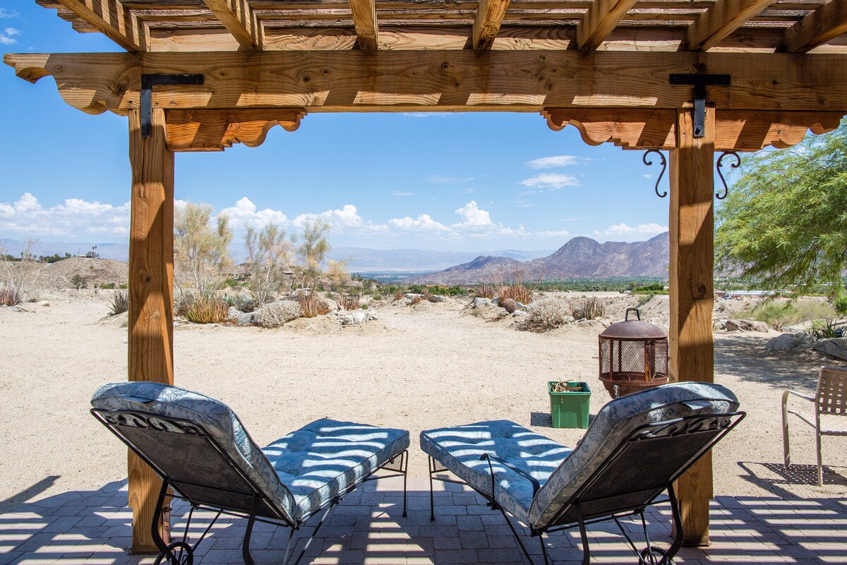 Two loungers are positioned under a wooden pergola, offering views of a desert landscape with mountains in the distance. A gentle breeze flows through the scene, highlighted by scattered desert vegetation. The sky features fluffy clouds and expansive blue hues.