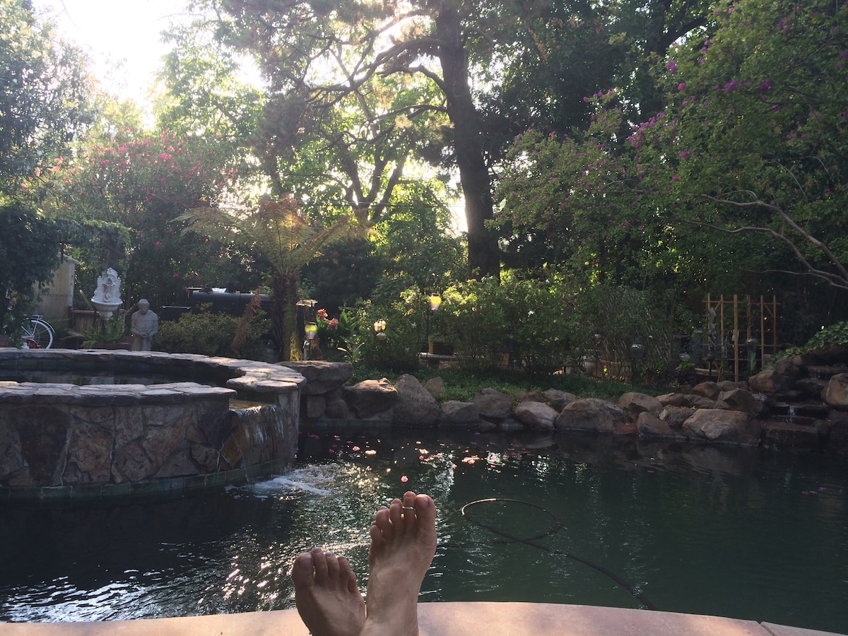 The backyard scene reveals lush greenery and vibrant flowers surrounding a tranquil pond. A stone waterfall gently flows into the water. Sunlight filters through trees, enhancing the serene environment. A stone patio edge is visible in the foreground, with bare feet resting beside the pond.