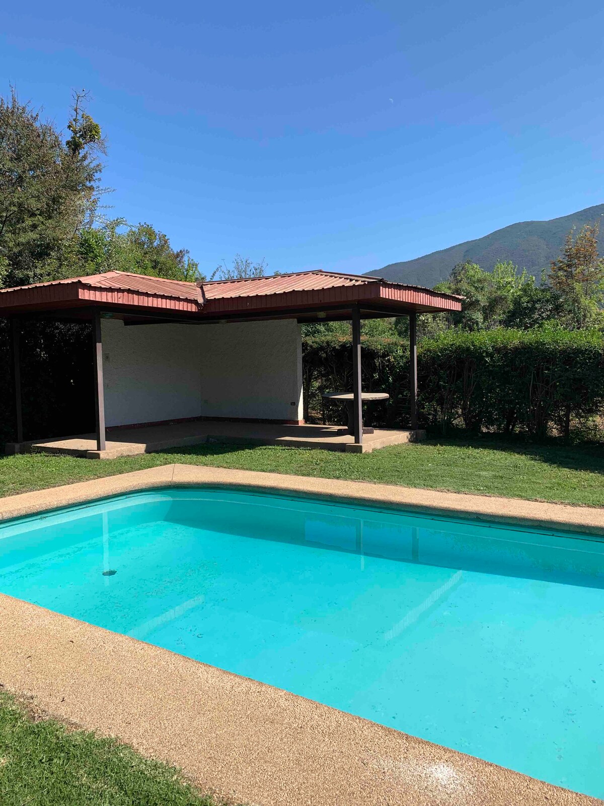 A clear blue swimming pool is framed by a well-maintained grassy area. A shaded structure with a tiled roof is visible nearby, offering outdoor seating. Lush greenery and a backdrop of distant mountains enhance the tranquil setting.