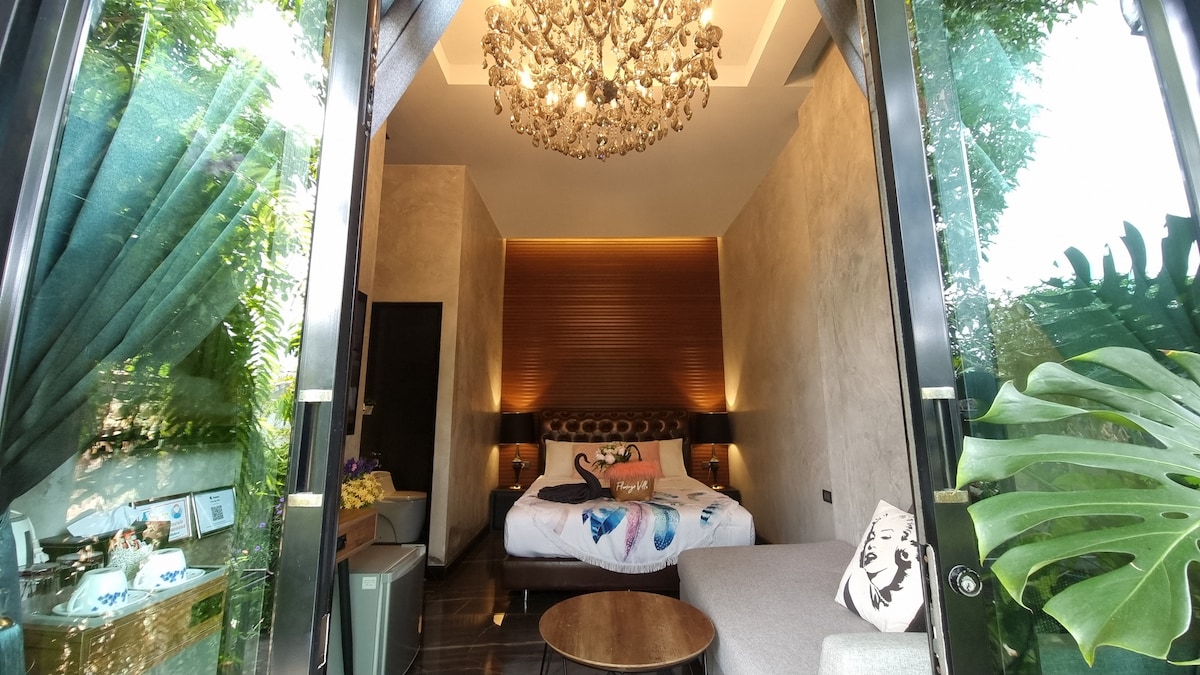 A modern bedroom features a spacious bed adorned with decorative pillows, complemented by a stylish chandelier overhead. Large glass doors open to a lush outdoor area, allowing natural light to flood the space. A comfortable sofa is situated nearby, enhancing the serene setting.