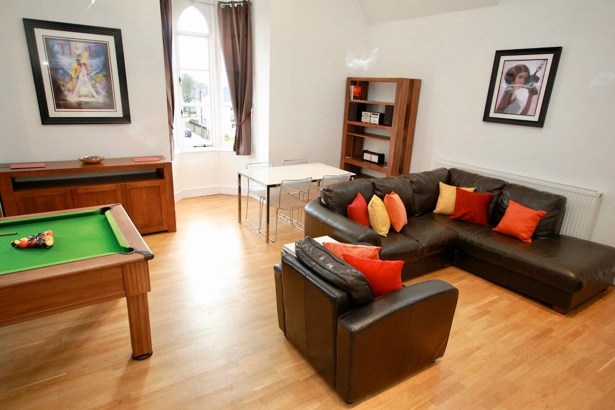 The spacious living room features a comfortable sectional sofa adorned with vibrant orange and black cushions. A pool table is positioned nearby, along with a glass dining table illuminated by natural light from arched windows, creating an inviting communal space.