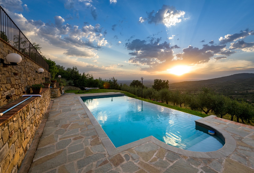 A serene outdoor pool is positioned against a backdrop of rolling hills and a dramatic sunset. Stone surfaces frame the pool area, while olive trees and lush greenery stretch into the distance, creating a tranquil atmosphere.