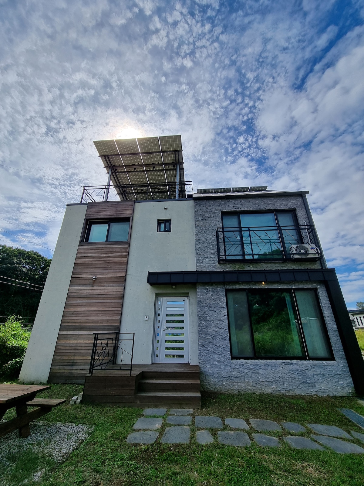 The exterior of the two-story house is showcased with large windows and a modern design. A deck with a railing is visible on the upper floor, and solar panels are installed on the roof. A pathway made of stepping stones leads to the entrance.