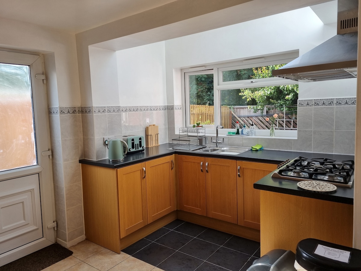 A well-equipped kitchen features wooden cabinetry and a black countertop. Natural light flows through a large window that overlooks a garden. A glass backsplash enhances the space, while kitchen appliances are neatly arranged, contributing to a functional and organized layout.