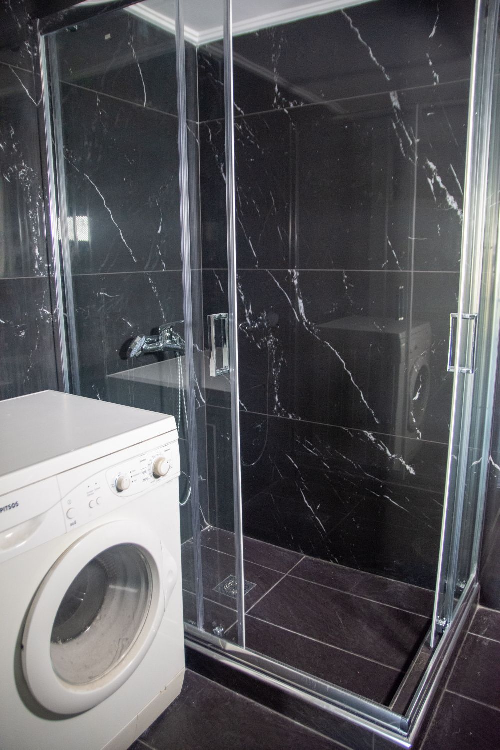 A sleek bathroom is presented with dark marble walls and a glass-enclosed shower. A washing machine is positioned next to the shower, featuring a simple design. The overall space is modern and functional, with contrasting light and dark surfaces.