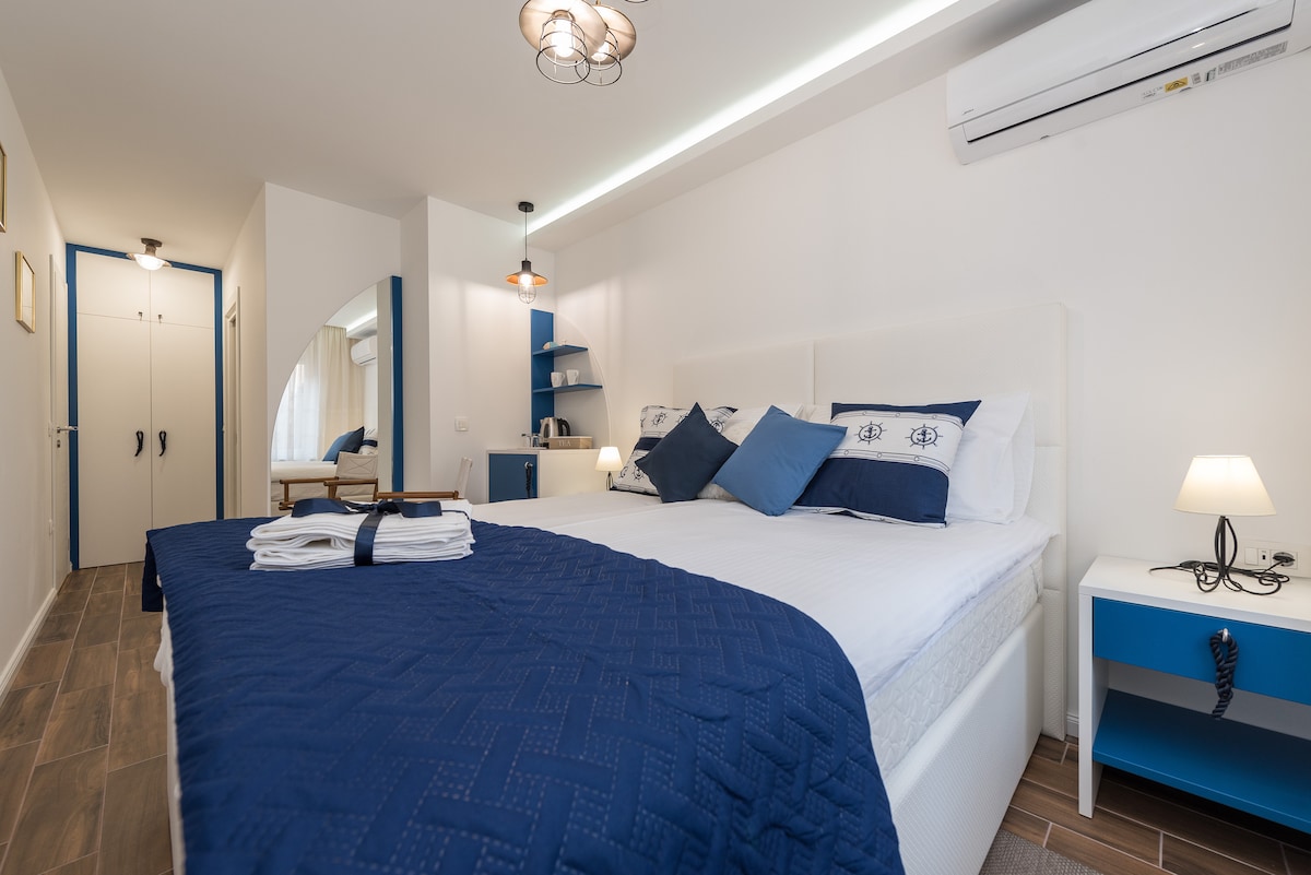 A modern double room is presented, featuring a spacious bed adorned with navy blue and white linens. A small seating area with light wood furniture is visible in the corner, complemented by storage cabinets. Air conditioning units and a soft glow from bedside lamps are included.