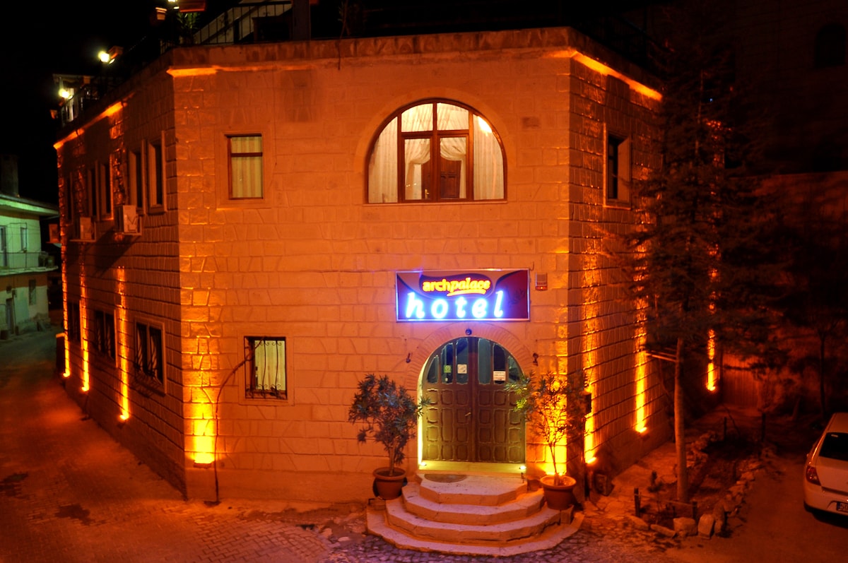 The hotel exterior is illuminated by warm lights at night, showcasing a stone facade with large arched windows. The entrance features a prominent sign displaying the name 'Arch Palace Hotel.' Potted plants are positioned at the doorway, enhancing the welcoming appearance.