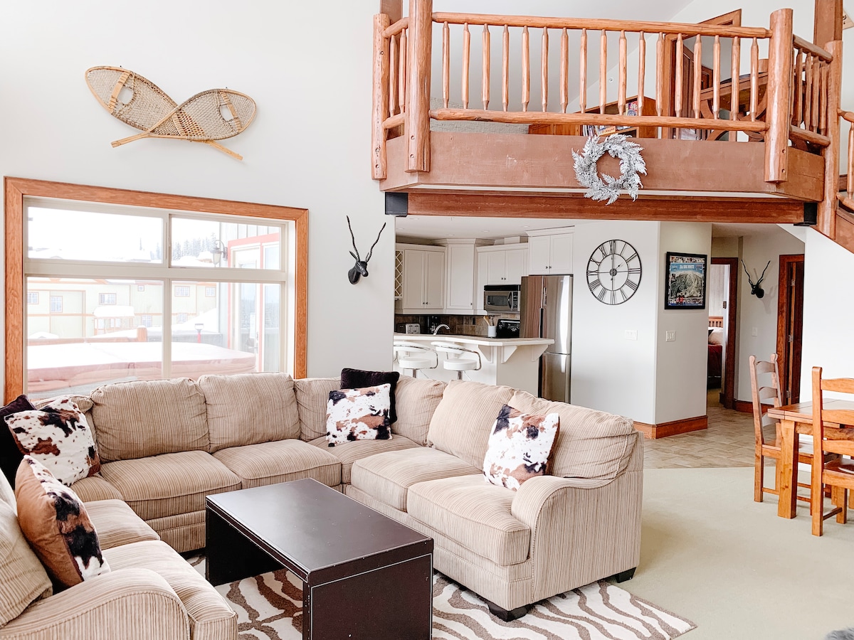 A spacious living area is highlighted by a large sectional sofa adorned with decorative cushions. A coffee table sits in front, and a patterned rug adds warmth to the space. Above, a lofted area adds vertical interest, while large windows provide ample natural light.