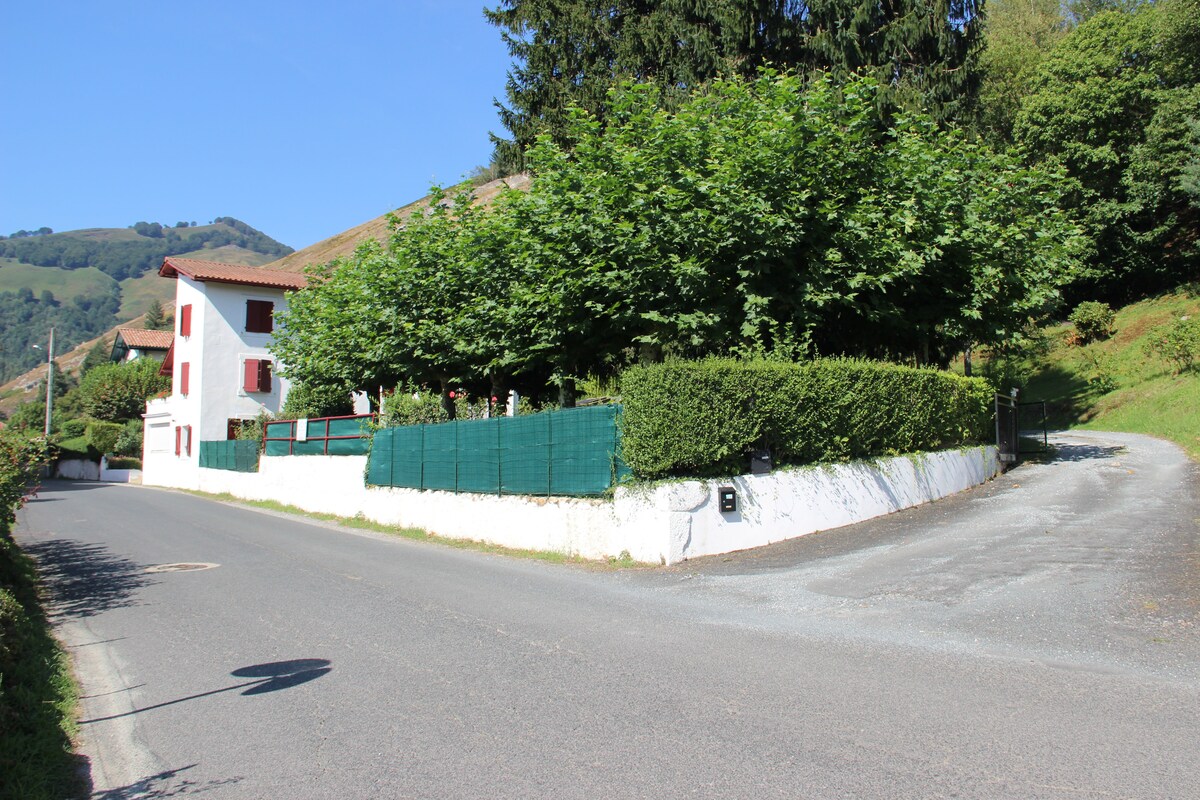 A two-story house with white exterior and red window shutters is situated at the curve of a quiet road. Lush green trees and neatly trimmed hedges surround the property, providing a natural barrier. A gravel path leads to the entrance, framed by gentle hillside landscape.