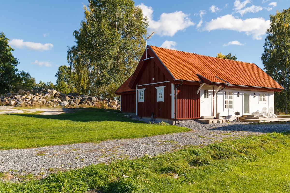 A charming red cottage features a distinctive roof and white trim, surrounded by a grassy area. A gravel pathway leads to the entrance. Large windows provide natural light, while a seating area with white chairs invites relaxation outdoors.