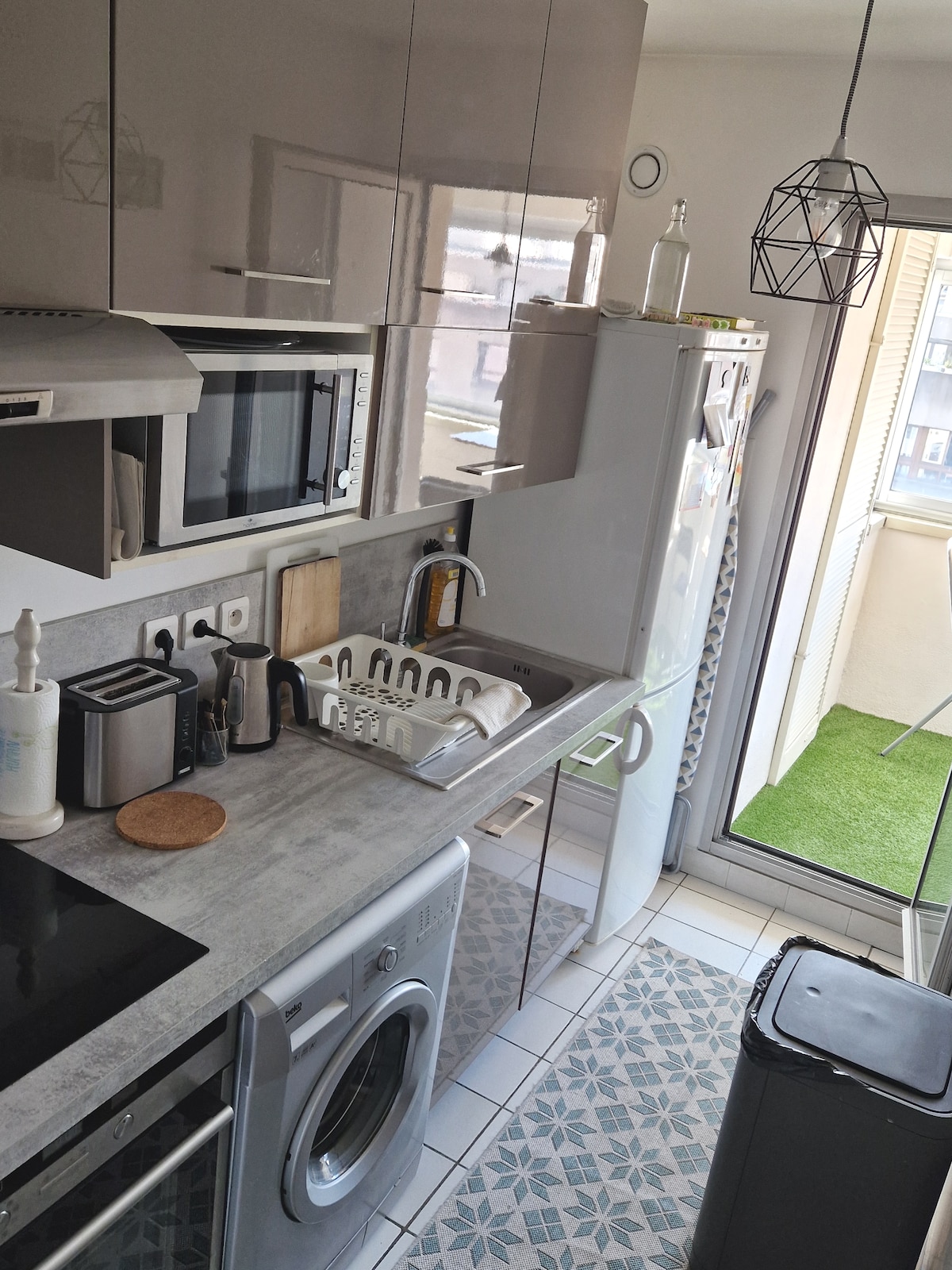 The kitchen features modern cabinetry and appliances, including a washing machine and microwave. Natural light enters through a window, illuminating the countertop and the tiled floor. A door leads to a small balcony with a grassy area, adding an outdoor touch.