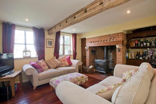 A comfortable lounge area features two plush sofas adorned with floral patterns. Exposed wooden beams are visible along the ceiling, while a brick fireplace adds charm. Natural light streams through the windows, illuminating shelves stocked with drinks and decorative items.