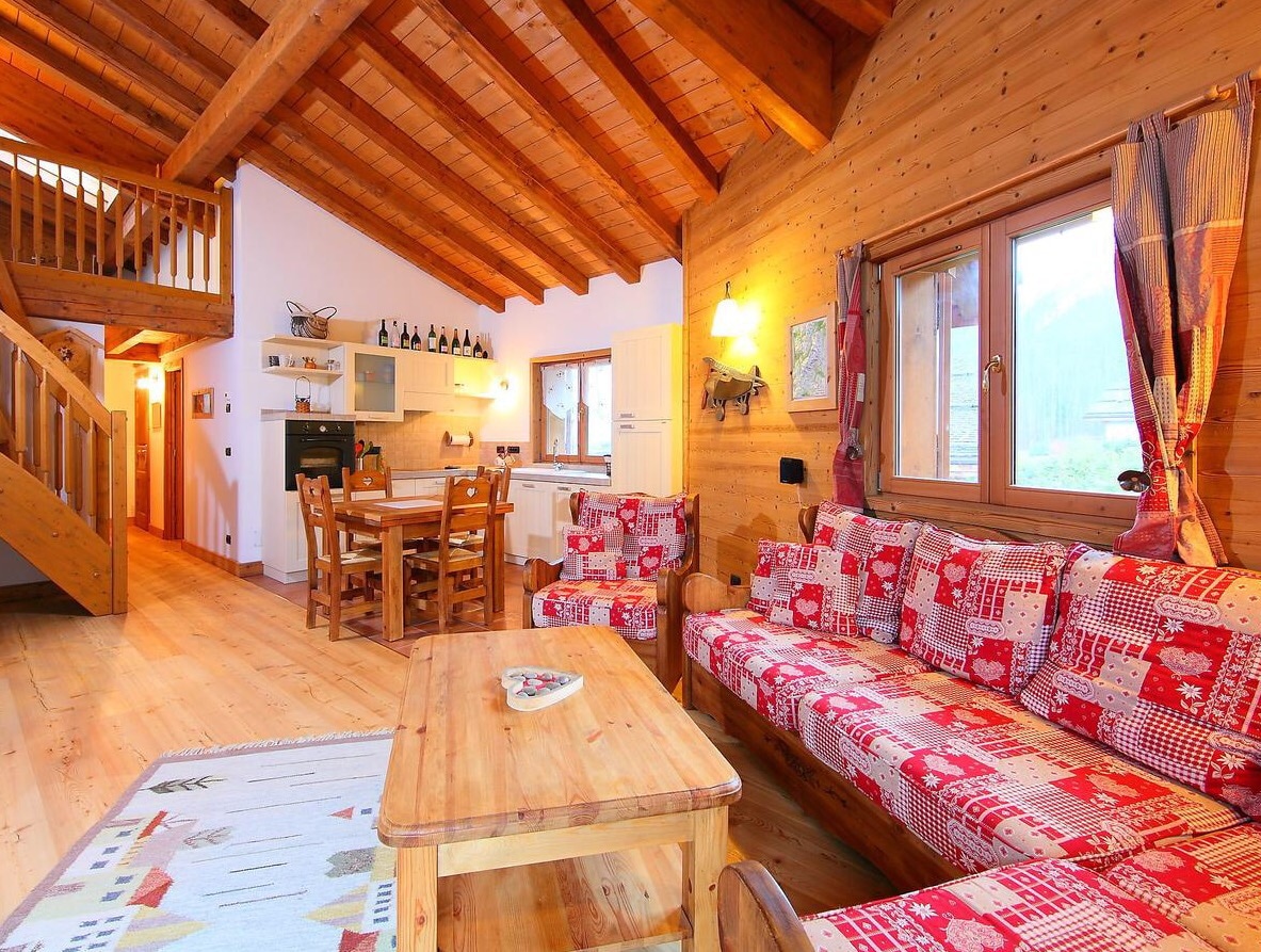 The living area features wooden beams and cozy decor. A comfortable seating arrangement is complemented by a wooden coffee table. A dining table for six is visible, along with a well-appointed kitchen area in the background. Natural light streams through large windows.