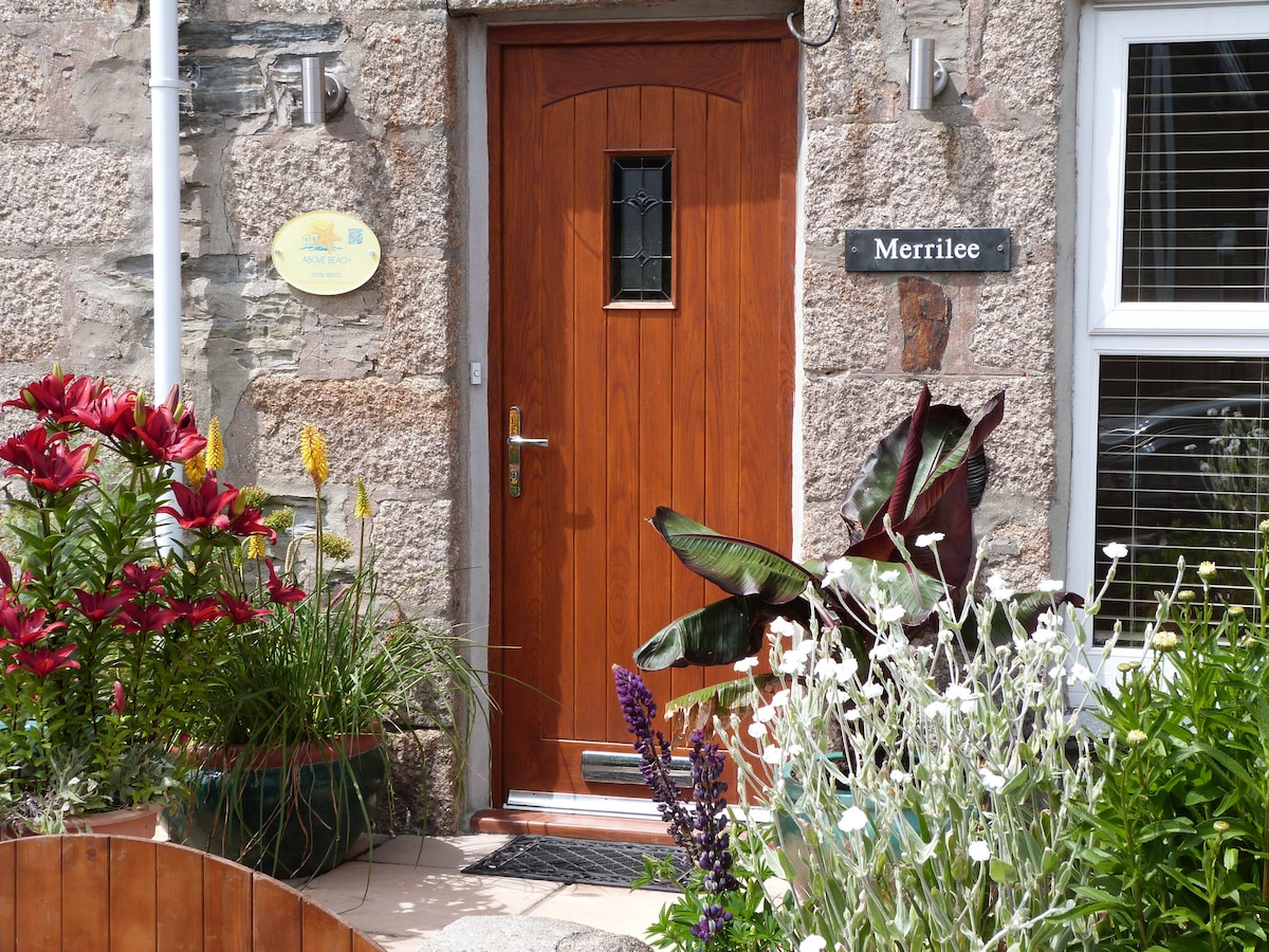 The entrance to Merrilee features a wooden door with a glass panel, surrounded by a stone wall. Colorful flowers in various planter boxes add vibrancy, while a sign indicates the name 'Merrilee.' Natural greenery provides a welcoming atmosphere.