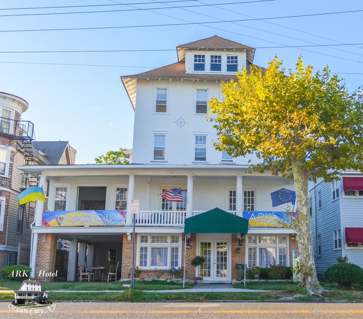 The exterior of the building displays a three-story structure with a flat roof and a covered entrance. Flags hang from the front, and a green awning provides shade. Surrounding trees offer greenery and enhance the welcoming atmosphere of the hotel.