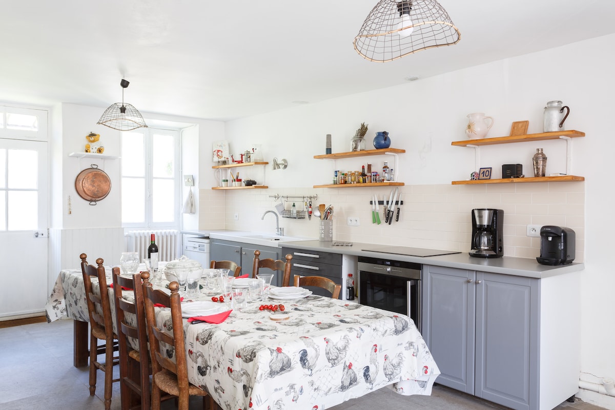 A spacious kitchen features a large wooden dining table set with plates and cutlery. Shelves display various kitchen items, while modern appliances blend with vintage decor. Natural light streams through windows, enhancing the room's functionality and inviting atmosphere.