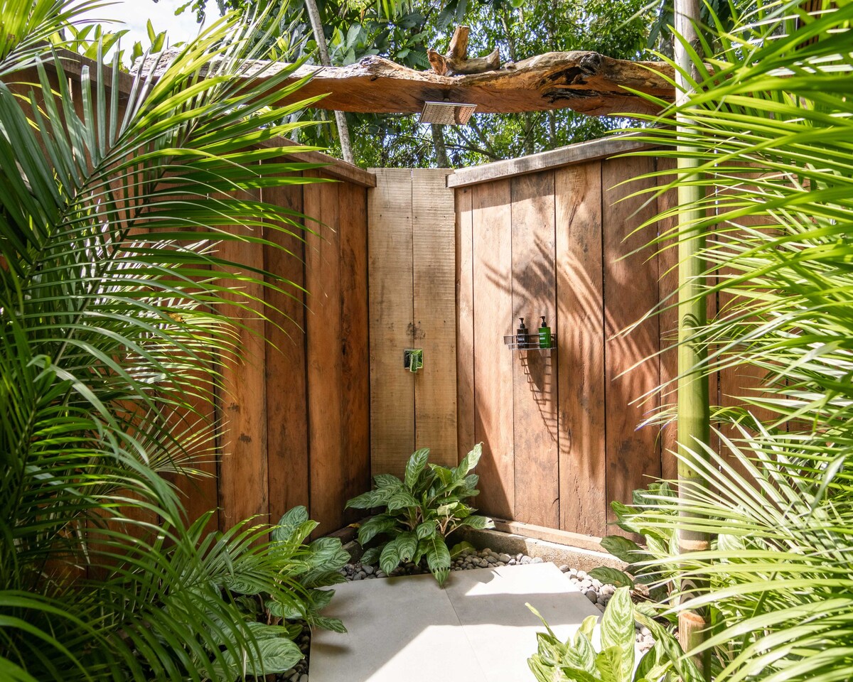 An outdoor shower area is surrounded by lush green foliage and wooden walls. The shower features natural elements, including a wooden bar overhead and a small shelf for toiletries. The stone flooring creates a serene space that integrates with the tropical environment.