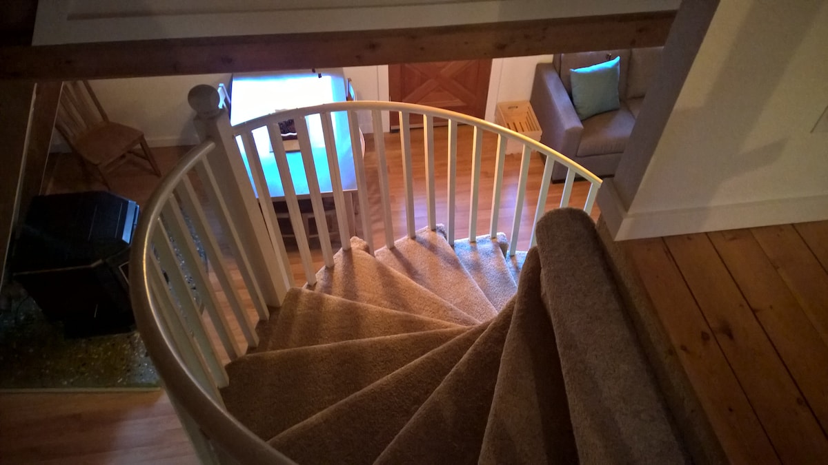 A spiral staircase is visible, showcasing a soft, carpeted path leading downward. The open living area can be seen below, featuring warm wooden floors and a cozy seating arrangement. Light subtly illuminates the space, enhancing the inviting layout of the chalet.