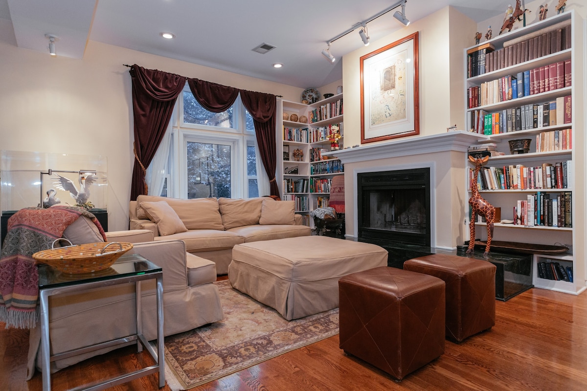 An inviting living area showcases a light-colored sectional sofa paired with a matching ottoman. A dark coffee table sits nearby, and a warm area rug lies underfoot. A bookshelf filled with books lines the wall, while soft lighting highlights the natural warmth of the wood flooring.