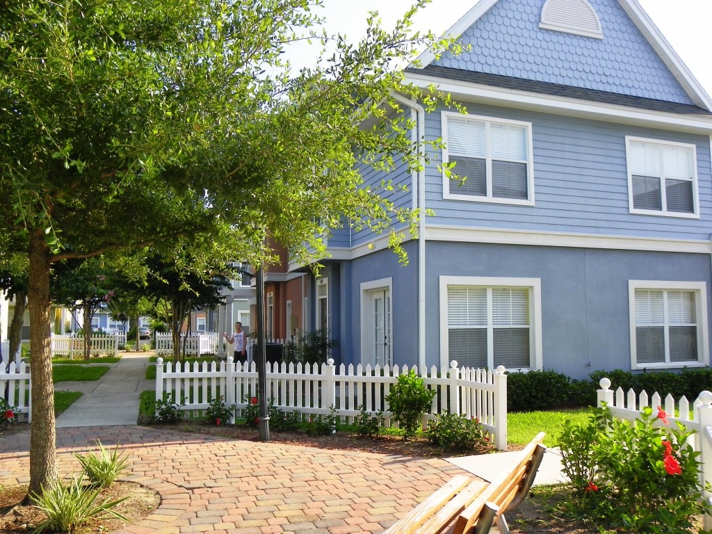 The exterior of a two-story townhouse is depicted, featuring a light blue facade and white picket fencing. Lush greenery surrounds the property, complemented by a brick pathway leading to the entrance. Natural sunlight enhances the vibrant colors of the landscaping.