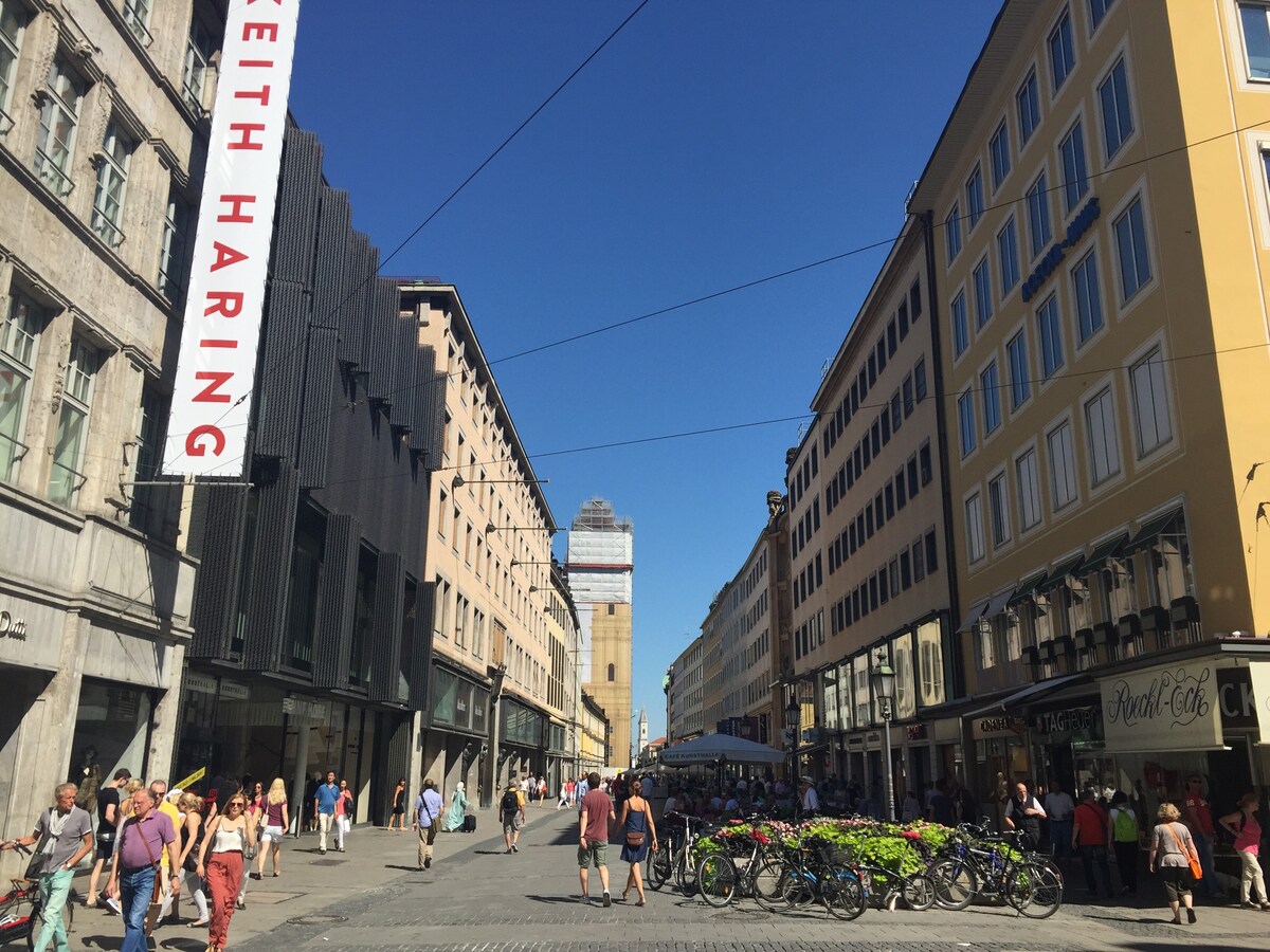 A bustling street scene in Munich features vibrant shops lining both sides, with pedestrians enjoying the sunny day. Colorful bicycles are parked along the curb, while an art banner hangs prominently. A historic building is visible in the distance, adding to the urban charm.