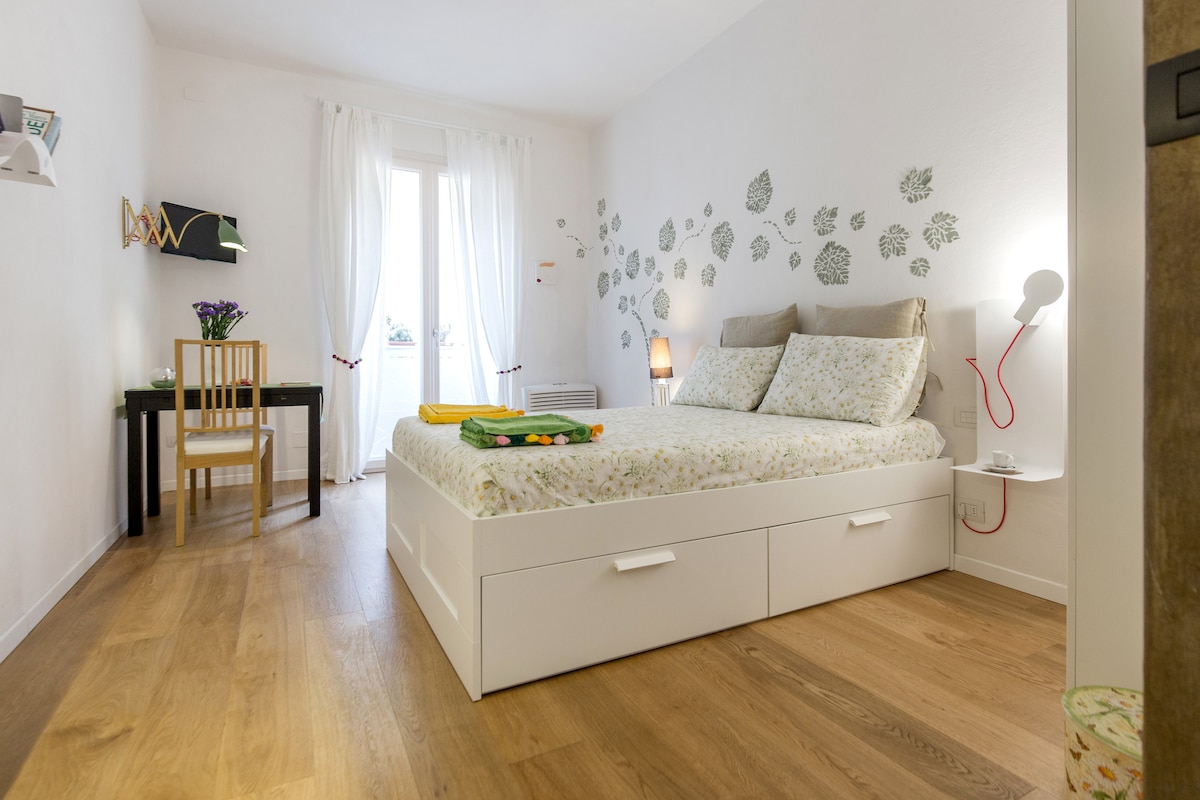 A bright studio room features light wooden flooring and a spacious bed with decorative bedding. A small table and chair set are positioned near the window, which is draped with sheer white curtains. Floral wall decals add a subtle touch of charm.