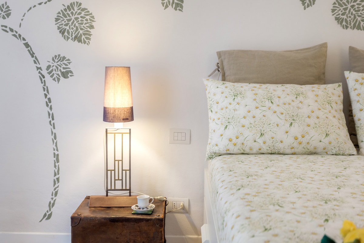 A well-lit corner of the bedroom features a spacious bed adorned with floral-patterned bedding. A lamp with a soft glow stands beside a small wooden stool, which holds a cup on a saucer. Decorative wall art with leaf motifs adds a touch of nature.