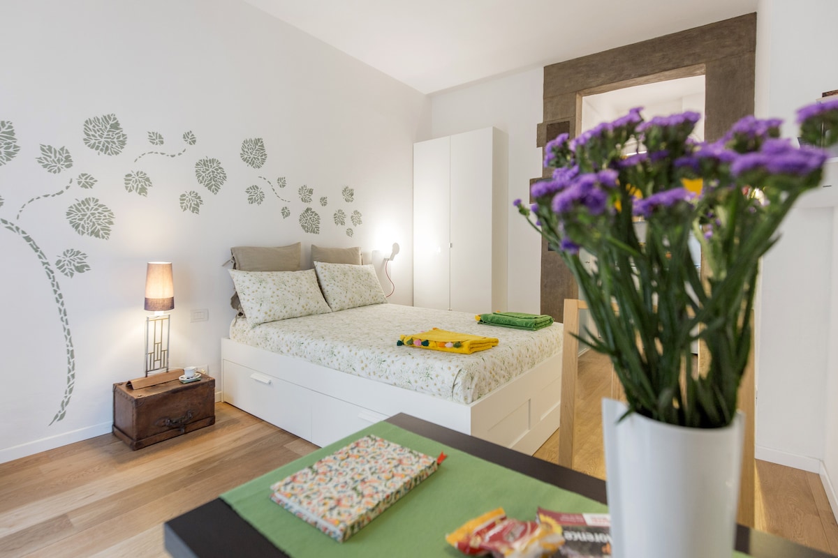 The studio features a bright sleeping area with a spacious bed, adorned in light floral patterns. A decorative wall with leaf motifs adds a natural touch. A small side table holds a green tablecloth and a vase with fresh flowers, enhancing the overall ambiance.