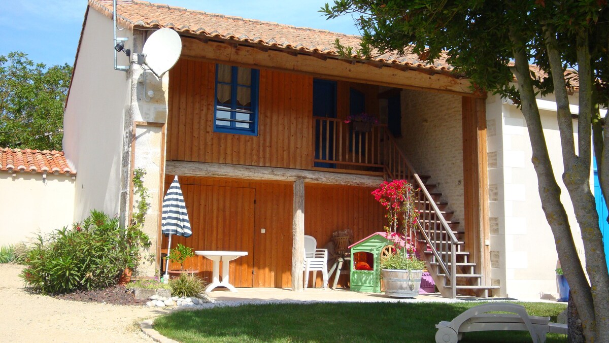 A two-story residence features a wooden balcony with blue shutters, overlooking a landscaped area. A staircase leads to the upper level, with a small white table and striped umbrella positioned nearby. Potted plants and colorful flowers add a vibrant touch to the outdoor setting.