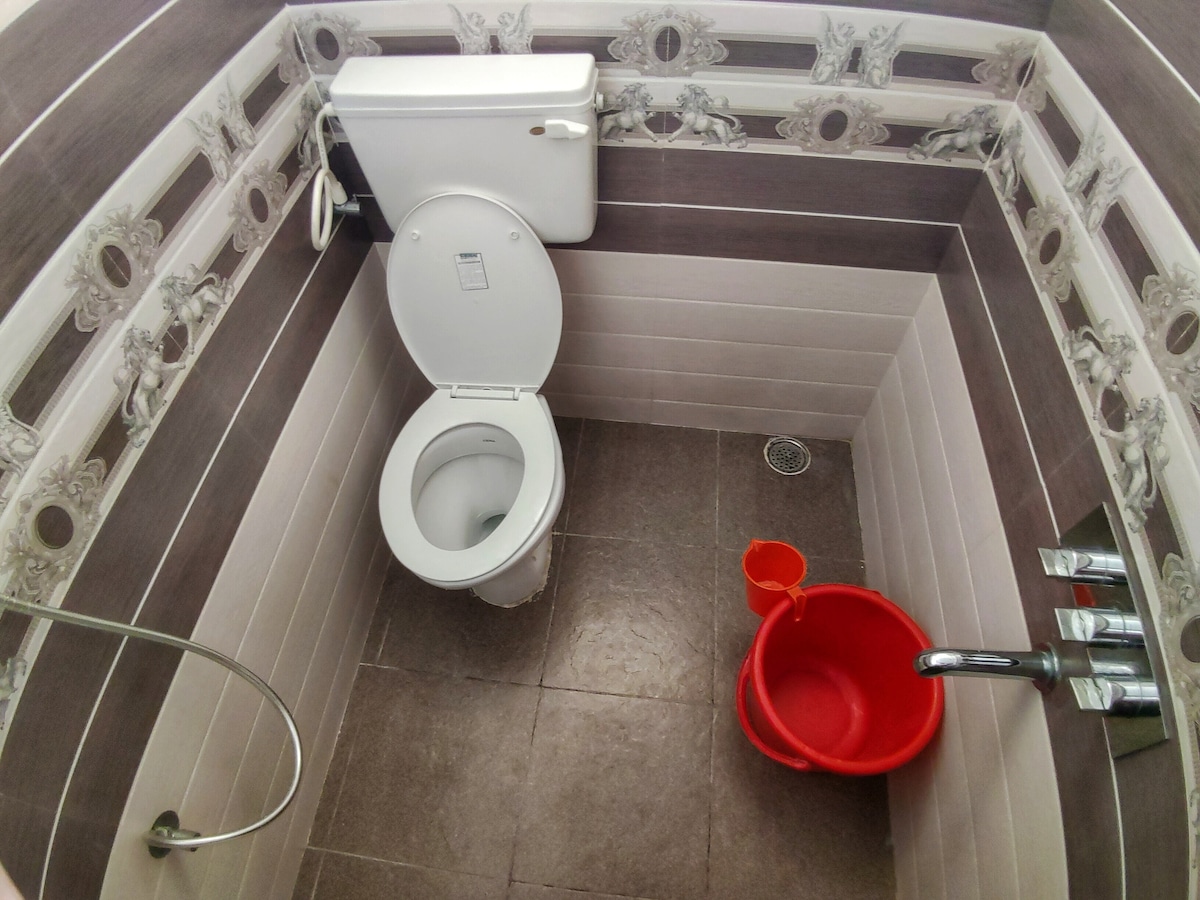 A well-maintained bathroom features a modern toilet, a shower area, and a red bucket for convenience. The walls are adorned with a mix of decorative tiles in subtle hues, while the flooring is a darker shade, providing a clean and functional space.