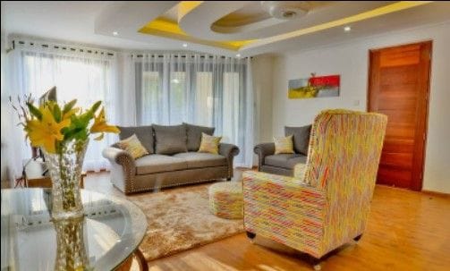 A bright living area features two stylish gray sofas and a colorful armchair, all arranged around a soft, textured rug. Large windows dressed with sheer curtains allow ample natural light to fill the space. A glass table reflects decorative flowers, enhancing the inviting atmosphere.