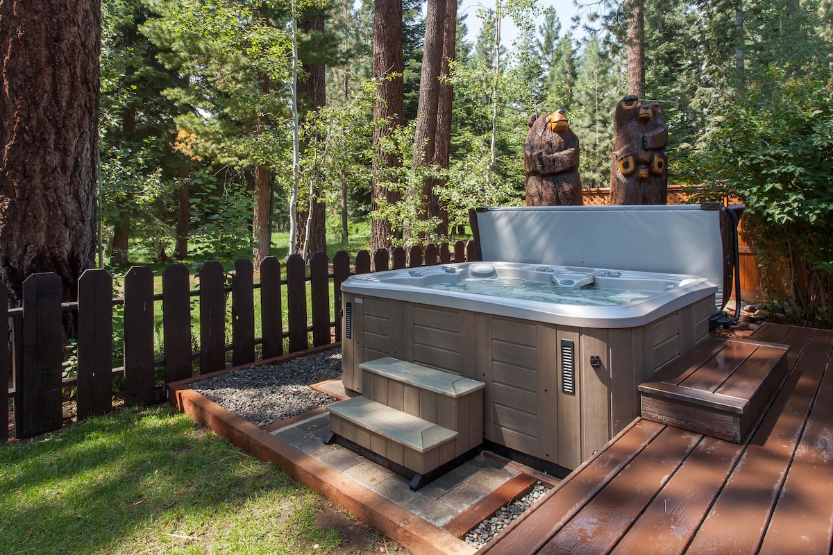 A hot tub is situated on a wooden deck surrounded by trees, providing a serene outdoor space. Steps lead up to the tub, landscaped with pebbles and grass for a natural feel. A wooden fence offers privacy while enhancing the tranquil atmosphere.