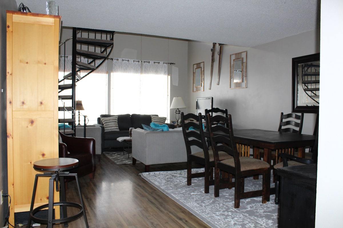 A spacious living and dining area is highlighted by a large dining table surrounded by black chairs. A cozy couch sits beside a window, with decorative mirrors and soft furnishings enhancing the space. A spiral staircase is visible, leading to an upper level.