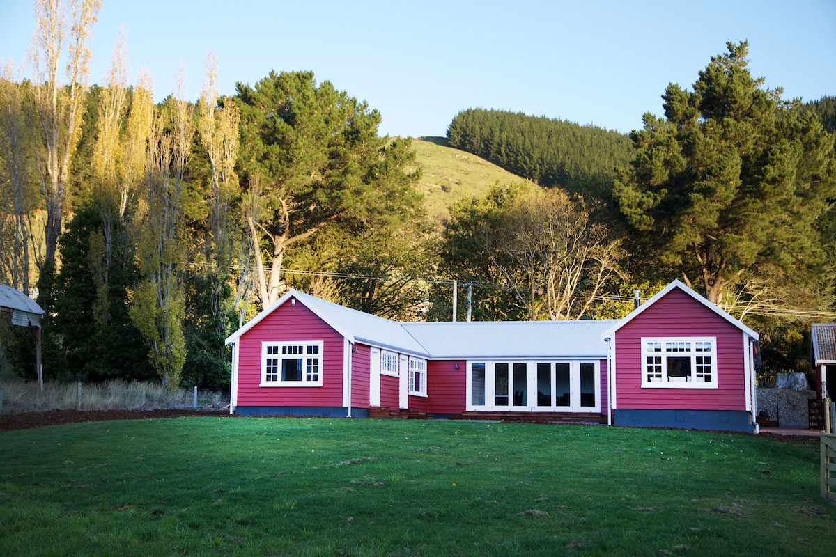 A charming barn-style building is shown, painted in a vibrant red with white trim. Large windows allow natural light to fill the interior, offering views of the expansive green lawn and surrounding hills. Tall trees frame the property, enhancing its serene outdoor setting.