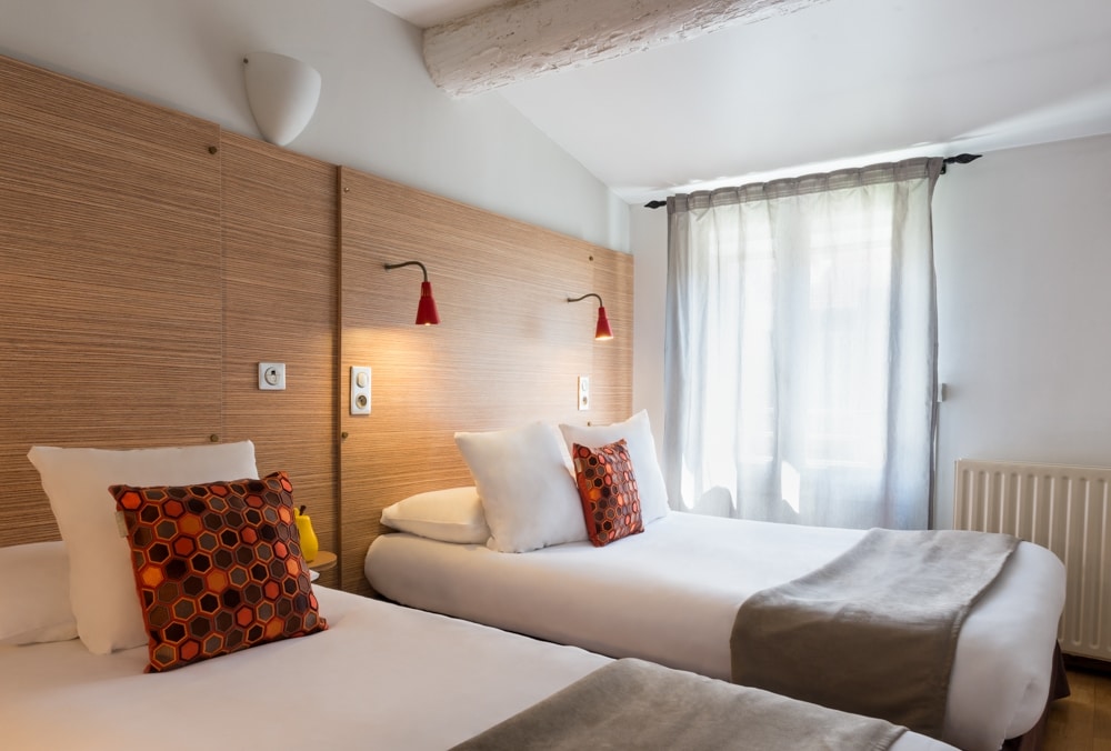 A room is presented with two beds, each adorned with decorative pillows. Natural light filters through a window with sheer curtains. The wood-paneled wall is complemented by modern light fixtures on either side of the beds, creating a functional yet cozy atmosphere.
