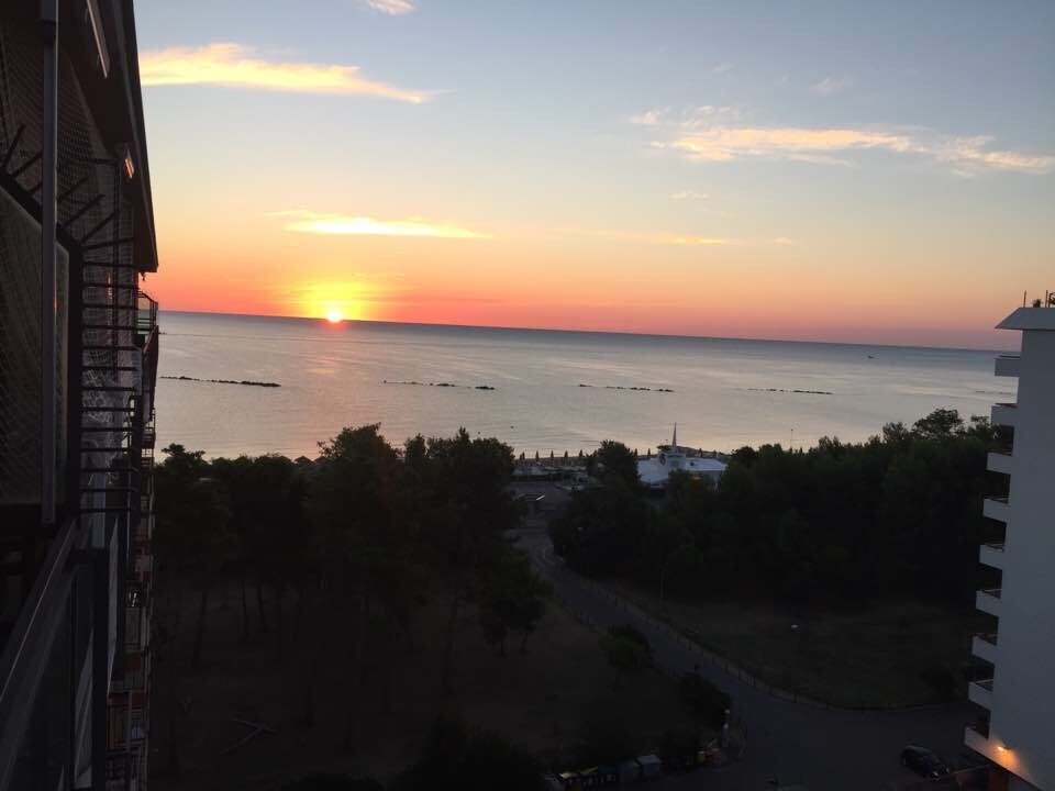 A stunning view of the sunset over the sea, with warm colors illuminating the sky. The horizon is visible, framed by gentle waves. In the foreground, trees and the outline of nearby buildings can be seen, enhancing the serene coastal atmosphere.