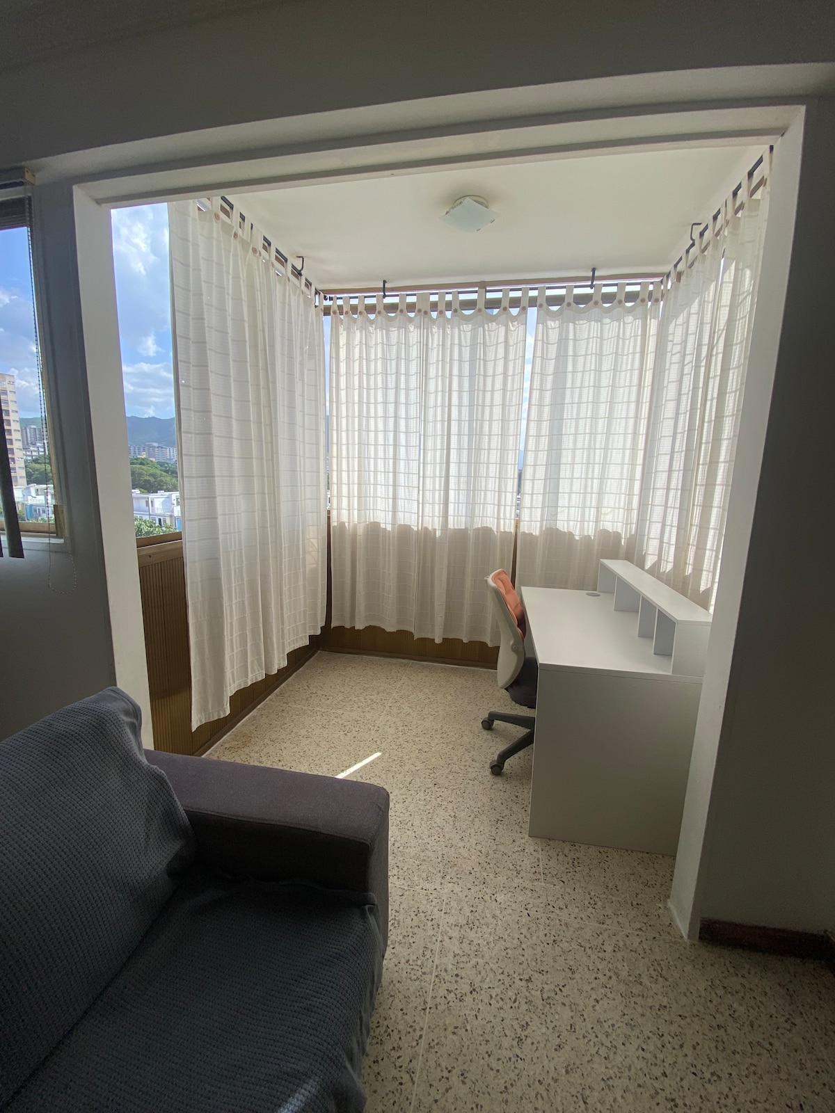 A cozy corner of the apartment is highlighted, featuring a workspace with a desk and chair, surrounded by sheer curtains that allow natural light to filter in. A comfortable sofa is visible, creating a welcoming atmosphere.
