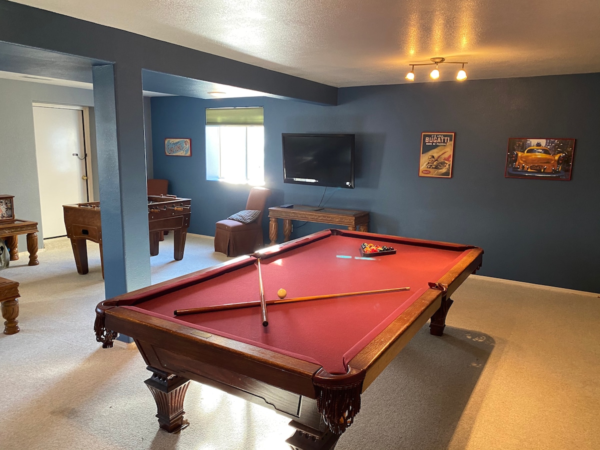 A spacious game room is highlighted by a red felt billiard table, equipped with cues and a set of balls ready for play. A flat-screen TV is mounted on the wall, while comfortable seating is nearby. Warm lighting enhances the inviting atmosphere.