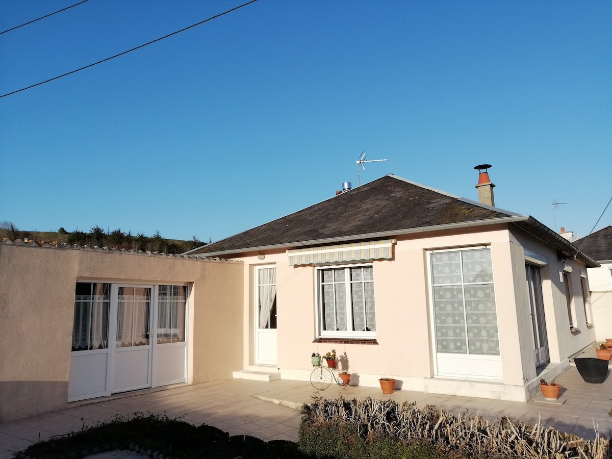 A single-story house is depicted, featuring a light-colored exterior with multiple windows. The front entrance is framed by a small porch area. Potted plants are visible along the base of the windows, and a clear blue sky provides a bright backdrop.