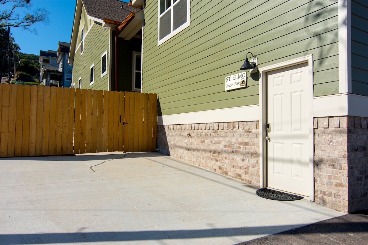 A private entrance is showcased, featuring a solid white door next to a wooden fence, with the 'St. Elmo' sign displayed above. The spacious parking pad is highlighted, offering a clear, unobstructed area for guest vehicles.