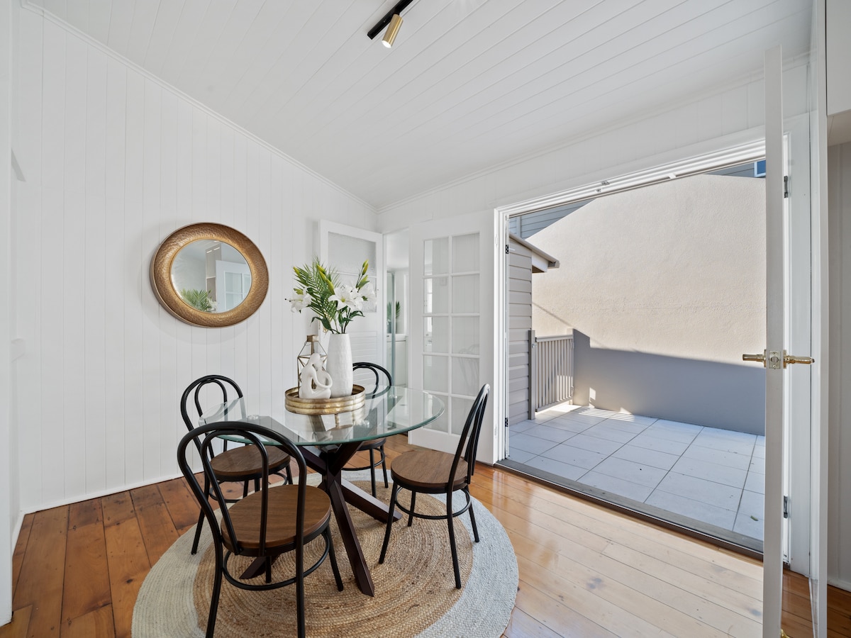 A dining area is showcased, featuring a round glass table surrounded by four wooden chairs. Natural light flows in through French doors that open to a patio, while a round mirror and a decorative vase with greenery enhance the space.