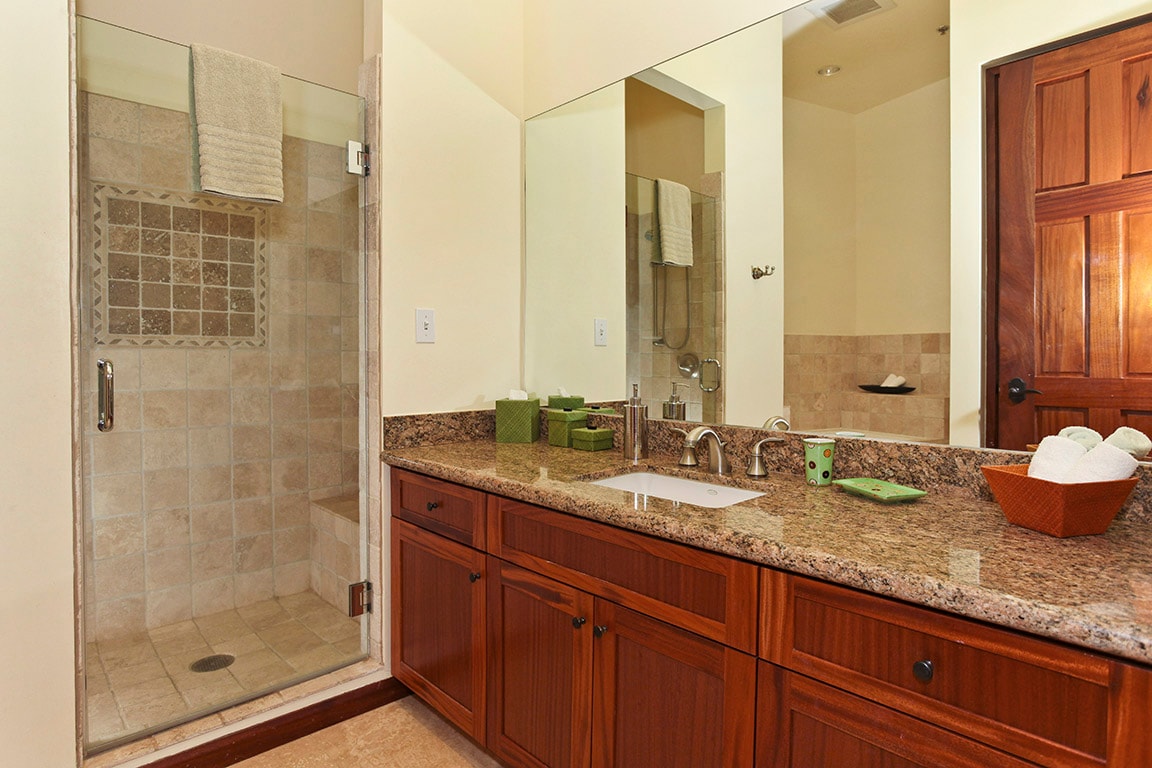 The bathroom features a walk-in shower with glass enclosure and tiled walls. A dual-sink vanity with dark wood cabinetry offers ample storage. Green accessories and towels provide a touch of color, while a large mirror enhances the room's brightness.