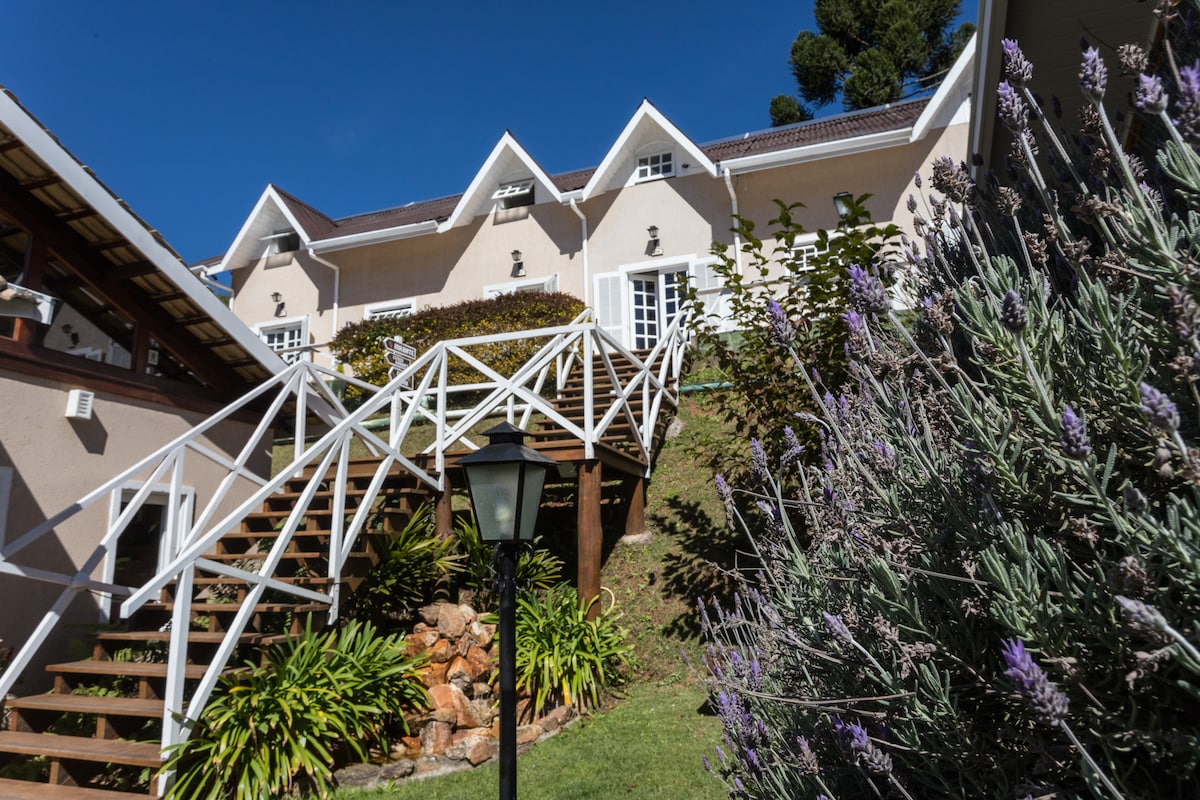 A charming exterior view showcases the building's sloped entrance and white railing leading up the stairs. Lush greenery surrounds the base, with lavender and other flowering plants offering muted color. Bright blue skies create a clear backdrop for the welcoming structure.