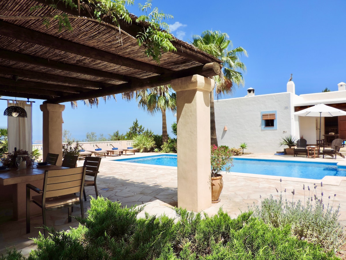 An outdoor area is presented with a covered patio featuring a dining table and chairs. A swimming pool is situated nearby, surrounded by greenery and sun loungers. The sky is clear, and palm trees add a tropical feel to the setting.