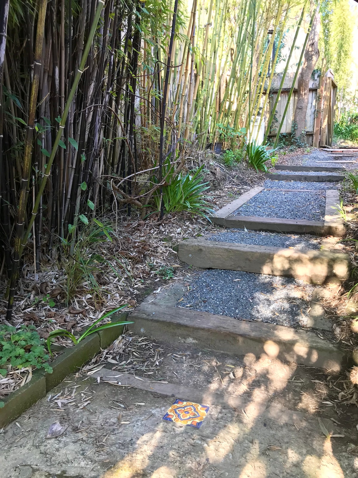 A stone pathway lined with natural foliage leads through a garden. The path, composed of wooden steps and gravel, meanders gently under the shade of tall bamboo. A decorative tile is visible near the bottom of the steps, adding a touch of color to the scene.