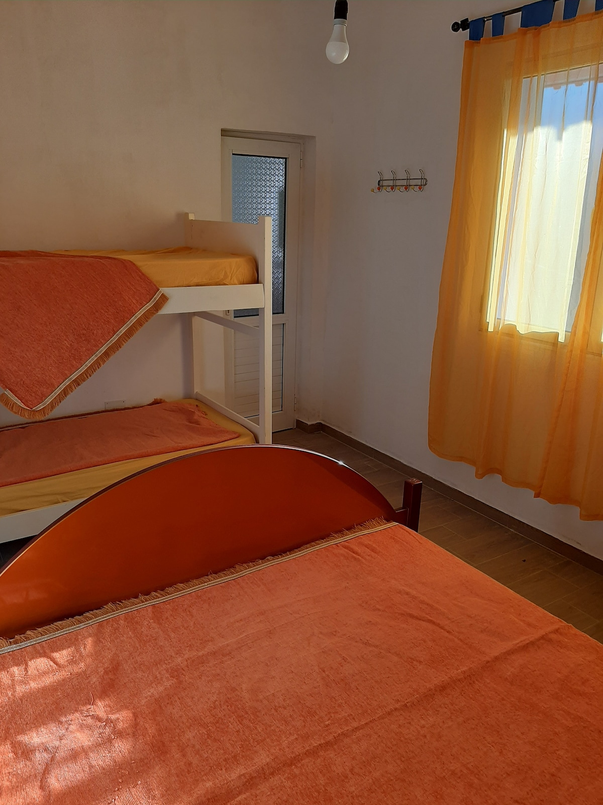 A cozy sleeping area is furnished with a spacious bed and a bunk bed arranged against the wall. Soft orange bedding complements the room's light-colored walls. Natural light filters in through a window dressed with sheer yellow curtains, enhancing the peaceful ambiance.