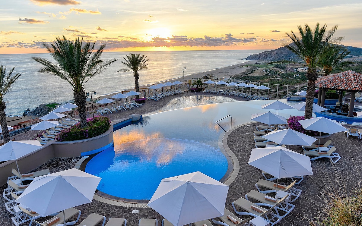 An expansive infinity pool is surrounded by lounge chairs and umbrellas, with palm trees casting gentle shadows. The sun sets over the horizon, casting warm hues across the sky and reflecting on the tranquil waters. The coastline is visible in the distance, adding to the serene atmosphere.